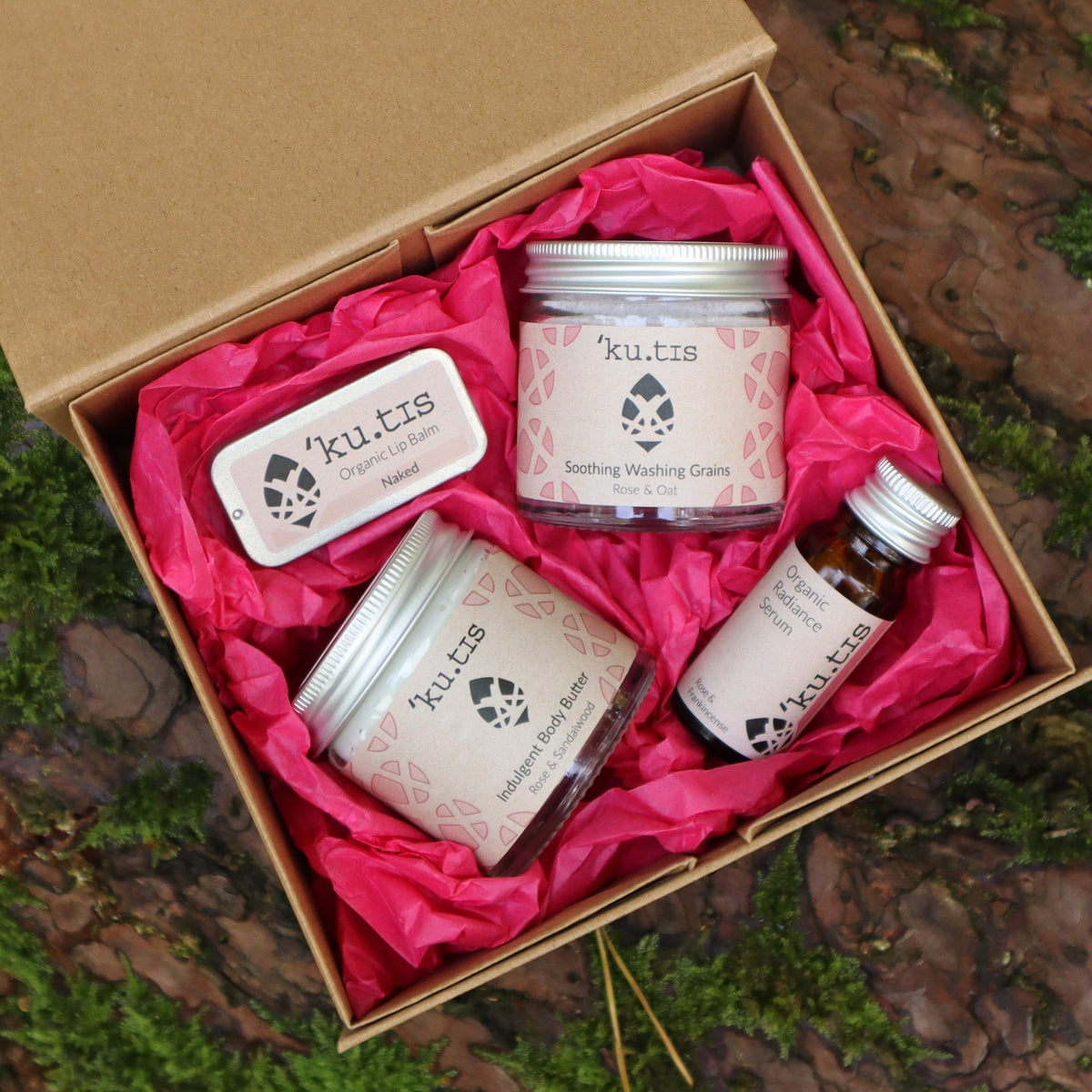 An open kraft gift box lined with pink tissue paper, containing naked lip balm, indulgent body butter, radiance serum and soothing washing grains. Made in Wales by Kutis Skincare.
