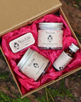 An open kraft gift box lined with pink tissue paper, containing naked lip balm, indulgent body butter, radiance serum and soothing washing grains. Made in Wales by Kutis Skincare.