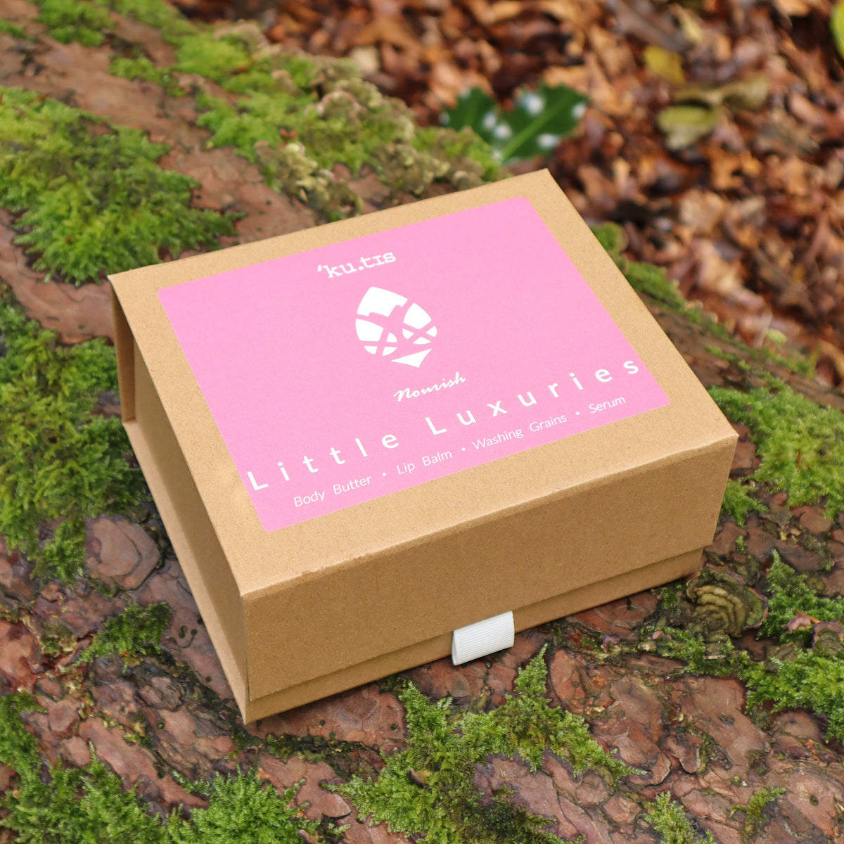 A kraft gift box with a pink label reading 'Little Luxuries' made by Kutis Skincare sits on a mossy log outdoors among fallen leaves and greenery.