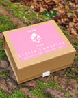 A kraft gift box with a pink label reading 'Little Luxuries' made by Kutis Skincare sits on a mossy log outdoors among fallen leaves and greenery.