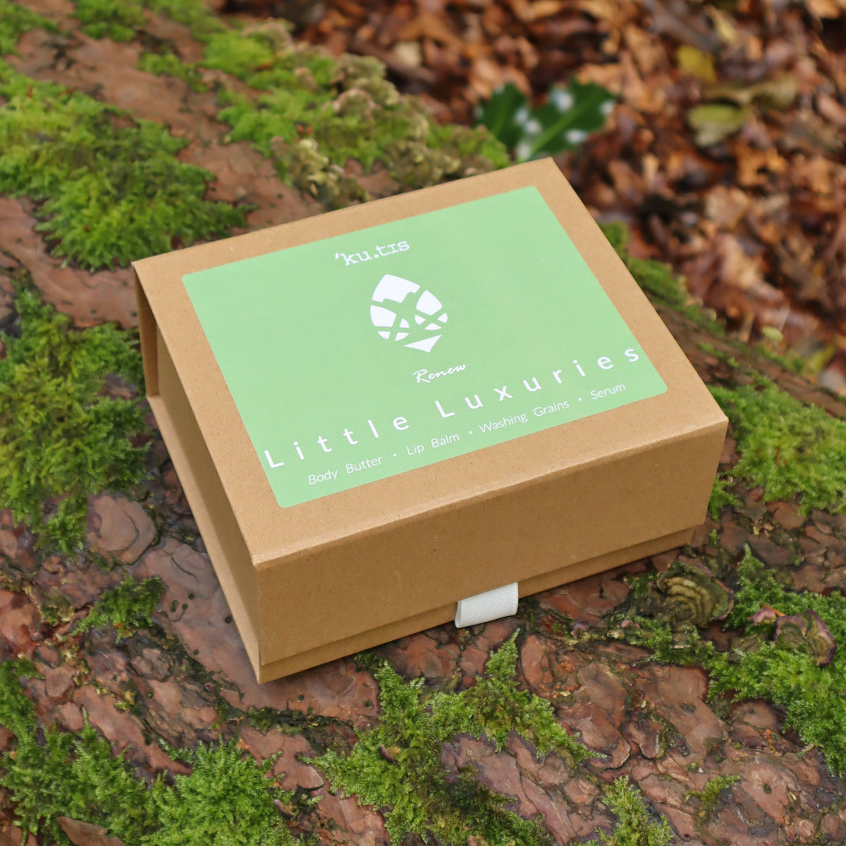 A square kraft paper gift box with a light green lid label reading 'Little Luxuries'  placed on a mossy log outdoors with forest ground and leaves blurred in the background. Made by Kutis Skincare