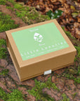 A square kraft paper gift box with a light green lid label reading 'Little Luxuries'  placed on a mossy log outdoors with forest ground and leaves blurred in the background. Made by Kutis Skincare