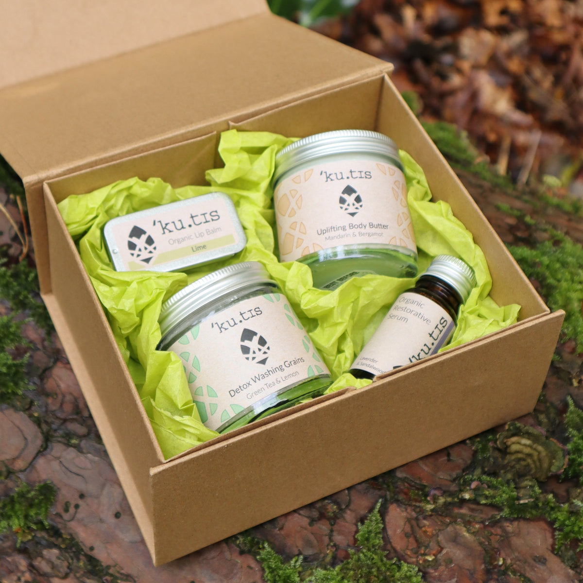 An open kraft gift box with a lime lip balm, uplifting body butter, Detox washing grains and restorative serum from the brand Kutis, placed on mossy ground  with fallen leaves in the background.
