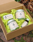 An open kraft gift box with a lime lip balm, uplifting body butter, Detox washing grains and restorative serum from the brand Kutis, placed on mossy ground  with fallen leaves in the background.